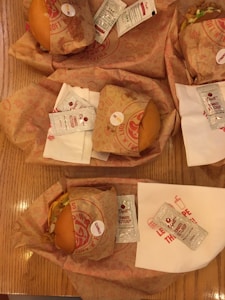 Several wrapped burgers are placed on a wooden table with packets of Dipt's tomato ketchup. Each burger is wrapped in branded paper, and white napkins are also visible beneath some of the packs.