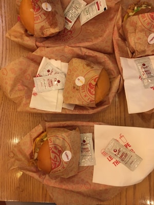Several wrapped burgers are placed on a wooden table with packets of Dipt's tomato ketchup. Each burger is wrapped in branded paper, and white napkins are also visible beneath some of the packs.