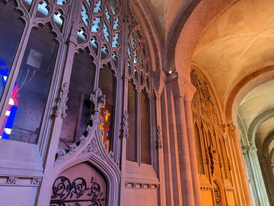 The image captures an ornate, gothic-style architectural structure with intricately carved stone arches and stained glass windows. The light filtering through the stained glass casts colorful reflections on the stone surfaces, creating a warm and vibrant atmosphere.
