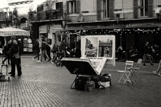 A busy street scene in a European city featuring an artist's table with paintings displayed. People walk by and some sit at outdoor café tables. The surroundings include classic architecture with a building that has balconies and shuttered windows.