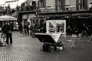 A busy street scene in a European city featuring an artist's table with paintings displayed. People walk by and some sit at outdoor café tables. The surroundings include classic architecture with a building that has balconies and shuttered windows.