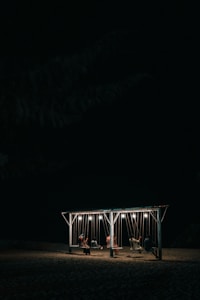 A group of people sit on swings attached to a wooden structure, illuminated by hanging lights in an otherwise dark setting. The ground appears sandy, suggesting a beach or outdoor location at night.
