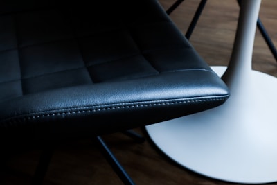 A close-up view of a dark leather chair seat beside a white, round table base. The chair has a smooth, cushioned surface with visible stitching. The table base is sleek and has a modern design. The background suggests a wooden floor.