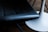 A close-up view of a dark leather chair seat beside a white, round table base. The chair has a smooth, cushioned surface with visible stitching. The table base is sleek and has a modern design. The background suggests a wooden floor.