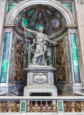 A marble statue depicting a robed figure in a dynamic pose, holding a long spear. The statue is set against a decorative backdrop featuring an arched niche with stained glass and marbled surfaces. Intricate architectural details and vibrant green and purple hues are present in the stained glass sections surrounding the statue.