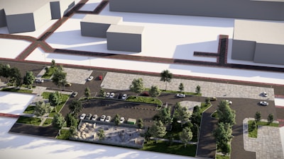 An aerial view of a modern urban parking lot surrounded by greenery. The parking area is organized with several cars parked neatly between rows of trees and pathways. Surrounding buildings appear as simplistic block structures.