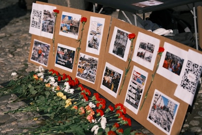 A display board is set up with several images and QR codes, each photo depicting scenes of destruction and aid efforts. Red carnations are pinned next to some of the images, and more flowers are laid at the base of the board.
