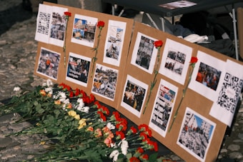 A display board is set up with several images and QR codes, each photo depicting scenes of destruction and aid efforts. Red carnations are pinned next to some of the images, and more flowers are laid at the base of the board.