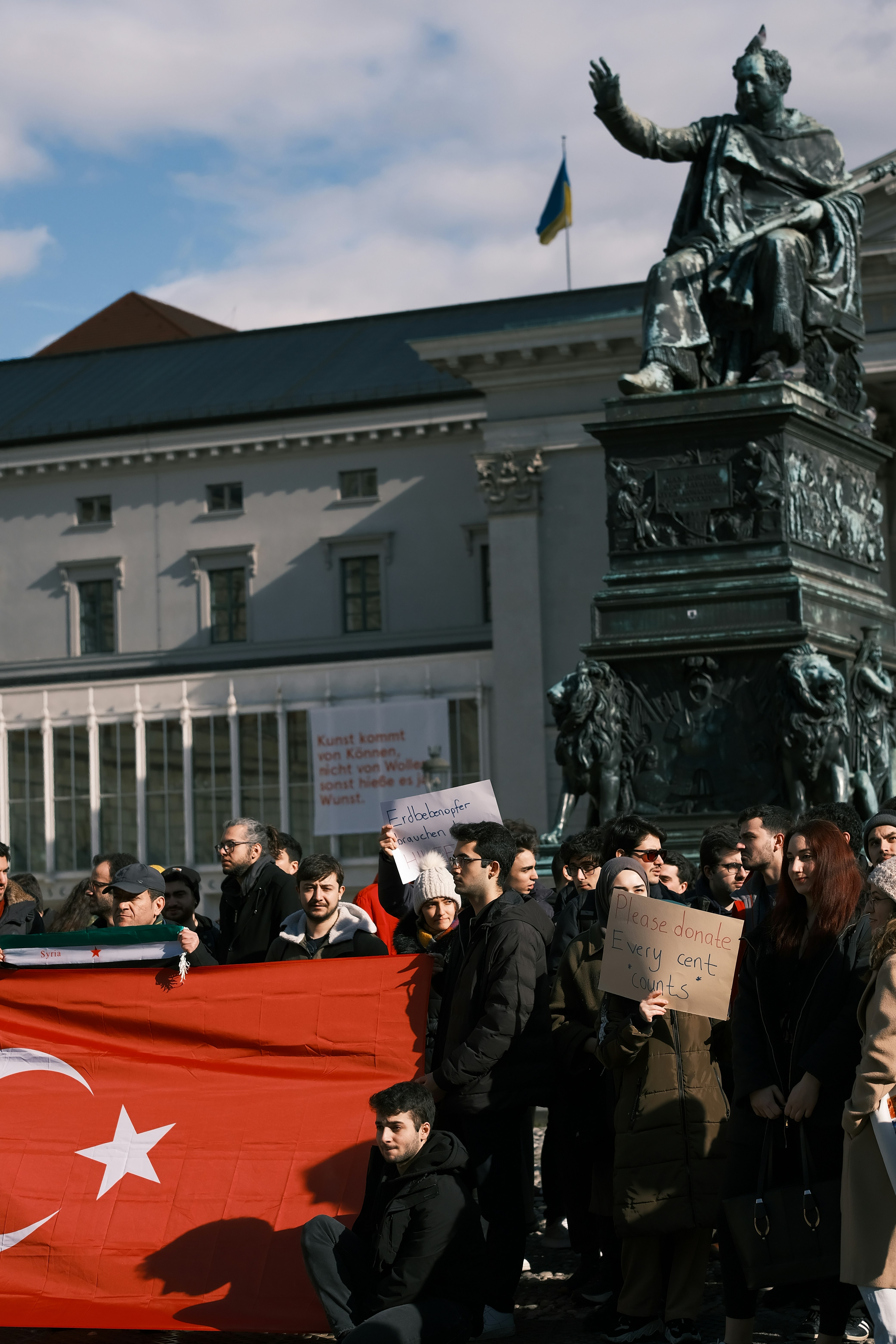 A group of people are gathered around a statue holding a large flag with a red background and white star and crescent. Some individuals hold signs with messages, one of which reads 'Please donate, every cent counts.' The setting includes a historical building in the background, and a blue and yellow flag is visible.