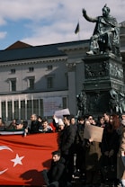 A group of people are gathered around a statue holding a large flag with a red background and white star and crescent. Some individuals hold signs with messages, one of which reads 'Please donate, every cent counts.' The setting includes a historical building in the background, and a blue and yellow flag is visible.