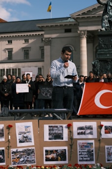 A man is holding a microphone and reading from a piece of paper in front of a crowd. The crowd behind him is holding up signs, and there's a large red flag with a white symbol on it. In the foreground, there is a board with images and red flowers attached to it. The scene appears to take place in front of a large, classical building with columns.