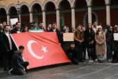 A group of people is gathered, holding a large Turkish flag. Some individuals are holding signs, including one that reads 'Please donate, every cent counts.' The setting features an architectural backdrop with arches.