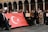 A group of people is gathered, holding a large Turkish flag. Some individuals are holding signs, including one that reads 'Please donate, every cent counts.' The setting features an architectural backdrop with arches.