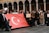 A group of people is gathered, holding a large Turkish flag. Some individuals are holding signs, including one that reads 'Please donate, every cent counts.' The setting features an architectural backdrop with arches.