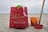 A red tote bag with white polka dots and German text sits on a sandy beach. Beside it, an orange plastic bucket adorned with starfish illustrations and a wooden shovel with a red spade are placed. The background shows an overcast sky with a few people barely visible in the distance.