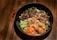 A black bowl containing grilled meat, sliced cucumbers, shredded carrots, crispy fried spring rolls, and garnished with herbs rests on a wooden surface.