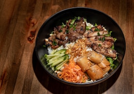 A black bowl containing grilled meat, sliced cucumbers, shredded carrots, crispy fried spring rolls, and garnished with herbs rests on a wooden surface.
