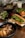 A wooden table is set with a variety of Vietnamese-style dishes. In the foreground, a black bowl contains grilled meat, likely pork, garnished with green onions and accompanied by crispy spring rolls and fresh bean sprouts. A crusty baguette sandwich filled with greens and shredded carrots is positioned alongside. In the background are additional bowls, one with a meat dish topped with greens, another with fresh herbs, and a glass of iced drink.