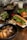 A wooden table is set with a variety of Vietnamese-style dishes. In the foreground, a black bowl contains grilled meat, likely pork, garnished with green onions and accompanied by crispy spring rolls and fresh bean sprouts. A crusty baguette sandwich filled with greens and shredded carrots is positioned alongside. In the background are additional bowls, one with a meat dish topped with greens, another with fresh herbs, and a glass of iced drink.