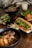 A wooden table is set with a variety of Vietnamese-style dishes. In the foreground, a black bowl contains grilled meat, likely pork, garnished with green onions and accompanied by crispy spring rolls and fresh bean sprouts. A crusty baguette sandwich filled with greens and shredded carrots is positioned alongside. In the background are additional bowls, one with a meat dish topped with greens, another with fresh herbs, and a glass of iced drink.