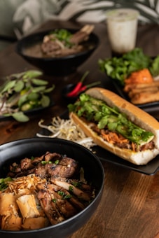 A wooden table is set with a variety of Vietnamese-style dishes. In the foreground, a black bowl contains grilled meat, likely pork, garnished with green onions and accompanied by crispy spring rolls and fresh bean sprouts. A crusty baguette sandwich filled with greens and shredded carrots is positioned alongside. In the background are additional bowls, one with a meat dish topped with greens, another with fresh herbs, and a glass of iced drink.