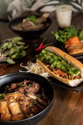 A wooden table is set with a variety of Vietnamese-style dishes. In the foreground, a black bowl contains grilled meat, likely pork, garnished with green onions and accompanied by crispy spring rolls and fresh bean sprouts. A crusty baguette sandwich filled with greens and shredded carrots is positioned alongside. In the background are additional bowls, one with a meat dish topped with greens, another with fresh herbs, and a glass of iced drink.