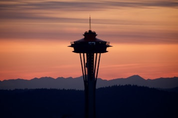 A silhouette of the Space Needle stands against a vibrant orange and pink sunset, with a backdrop of mountains and layered clouds creating a dramatic and peaceful scene.