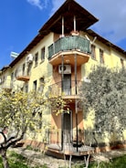 A multi-story building with yellow and peach-colored walls features several ornate balconies with wrought iron railings. There are air conditioning units mounted on the walls and a solar water heater on the roof. In the foreground, a tree with yellow fruits adds a natural element to the scene.