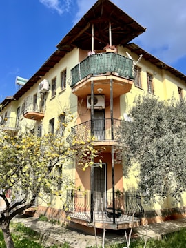 A multi-story building with yellow and peach-colored walls features several ornate balconies with wrought iron railings. There are air conditioning units mounted on the walls and a solar water heater on the roof. In the foreground, a tree with yellow fruits adds a natural element to the scene.
