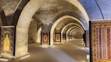 A series of intricately designed rugs are displayed in a stone archway corridor with soft lighting. The arches have a historic architectural style, suggesting an ancient or traditional setting. The corridor is lined with upright frames holding each rug, showcasing their vibrant patterns and colors.