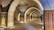 A series of intricately designed rugs are displayed in a stone archway corridor with soft lighting. The arches have a historic architectural style, suggesting an ancient or traditional setting. The corridor is lined with upright frames holding each rug, showcasing their vibrant patterns and colors.