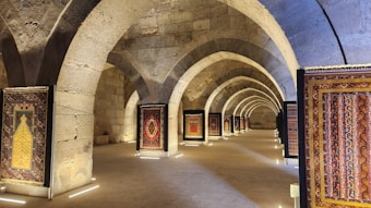 A series of intricately designed rugs are displayed in a stone archway corridor with soft lighting. The arches have a historic architectural style, suggesting an ancient or traditional setting. The corridor is lined with upright frames holding each rug, showcasing their vibrant patterns and colors.