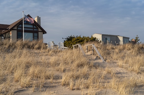 A scenic beachside house with large windows sits atop a sandy dune covered in dried grass. An American flag is displayed on a flagpole next to the house. The sky has a subtle overcast, suggesting a calm atmosphere with soft lighting. Two white wooden chairs are placed near the building, providing a place to sit and enjoy the view.