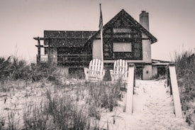 A two-story house with a modern design stands in a snowy landscape. There are two Adirondack chairs positioned in front, surrounded by tall grass on sandy ground. An American flag is visible on a pole by the house.