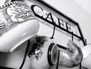 Several mugs with intricate black and white designs hang on hooks attached to a metal frame with the word 'CAFE' inscribed on it.