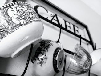 Several mugs with intricate black and white designs hang on hooks attached to a metal frame with the word 'CAFE' inscribed on it.