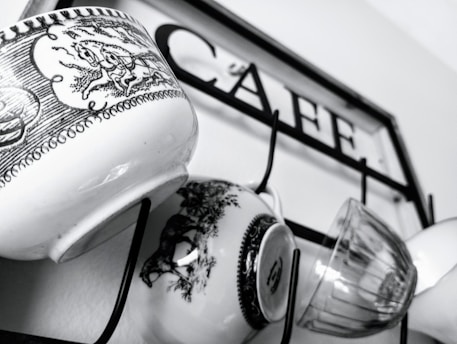 Several mugs with intricate black and white designs hang on hooks attached to a metal frame with the word 'CAFE' inscribed on it.