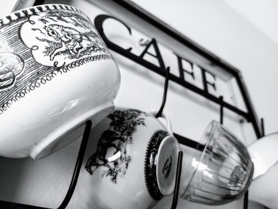 Several mugs with intricate black and white designs hang on hooks attached to a metal frame with the word 'CAFE' inscribed on it.