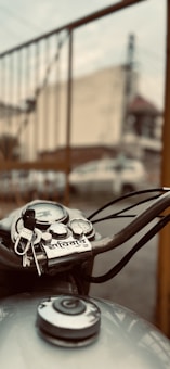 A close-up of a motorcycle's handlebar featuring a set of keys resting on it. The background is blurred, showcasing a fence and some buildings. The focus is on the metallic and mechanical elements of the motorcycle with some text visible near the handlebars.