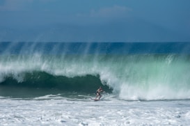 A surfer is skillfully navigating a large wave, with mist and spray filling the air. The ocean's hues range from deep blue to vibrant green, with the sunlight creating a shimmering effect on the water's surface. The image captures the dynamic and powerful movement of the wave and the surfer's concentration and balance.
