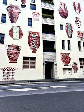 A building facade adorned with large, decorative murals of ancient vases. These vases feature intricate patterns and designs in red and white colors. Windows are interspersed among the murals. The building's entrance is labeled 'Excelsior' and is bordered by simple geometric patterns. A small sidewalk runs parallel to the street in front of the building.