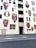 A building facade adorned with large, decorative murals of ancient vases. These vases feature intricate patterns and designs in red and white colors. Windows are interspersed among the murals. The building's entrance is labeled 'Excelsior' and is bordered by simple geometric patterns. A small sidewalk runs parallel to the street in front of the building.