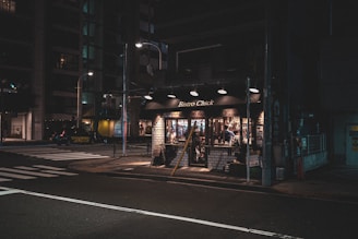 A dimly lit street corner featuring a small, cozy bistro with the sign 'Bistro Chick'. The bistro has brick walls and large windows with warm lighting inside, creating a welcoming atmosphere. There are a few people inside, visible through the glass, engaged in conversation. The surrounding street is quiet, with crosswalk lines and a few parked cars.