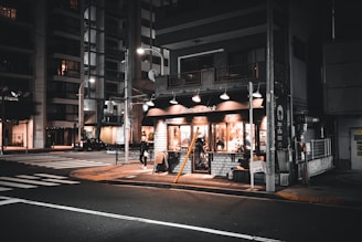 A cozy corner bistro with warm lighting emanating from its windows during nighttime. The street is quiet and dimly lit, with tall buildings visible in the background. A lone pedestrian walks past the bistro, adding a sense of calm and solitary atmosphere.