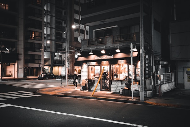 A cozy corner bistro with warm lighting emanating from its windows during nighttime. The street is quiet and dimly lit, with tall buildings visible in the background. A lone pedestrian walks past the bistro, adding a sense of calm and solitary atmosphere.