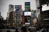 A bustling urban scene with numerous high-rise buildings featuring large colorful advertisements and billboards. The streets are crowded with people and vehicles, suggesting a busy city intersection. Various brands and businesses, like Starbucks, are visible, and digital screens display animated content.