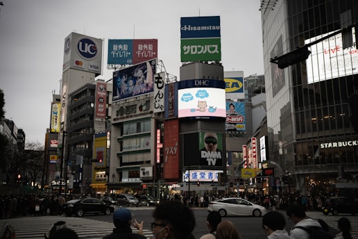 A bustling urban scene with numerous high-rise buildings featuring large colorful advertisements and billboards. The streets are crowded with people and vehicles, suggesting a busy city intersection. Various brands and businesses, like Starbucks, are visible, and digital screens display animated content.