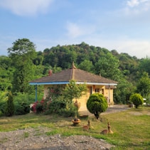 A small rural house with a tiled roof is surrounded by lush greenery and colorful flowers. The grass in the foreground appears well-maintained, and several shrubs are strategically planted around the area. The surrounding trees are dense, creating a backdrop of natural beauty under a blue sky.