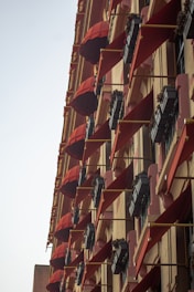 A tall building facade adorned with multiple red awnings and flower boxes mounted on the exterior. Each window has a triangular red canopy extending outward, providing shade and a decorative element. The flower boxes underneath are filled with small green plants.