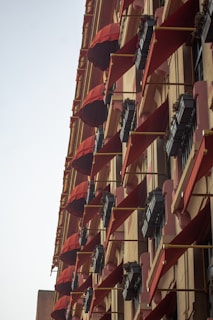 A tall building facade adorned with multiple red awnings and flower boxes mounted on the exterior. Each window has a triangular red canopy extending outward, providing shade and a decorative element. The flower boxes underneath are filled with small green plants.